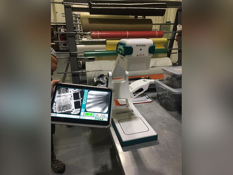 Image in a manufacturing environment with rolls of material on a stand in the background. In the foregrond is table with the finished portable X-ray device. A person's hand is holding a tablet, showing an x-ray image of hand as part of a demonstration in RWC's Utah facility.